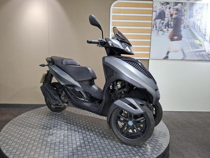 A gray three-wheeled scooter displayed on a circular metal platform, with a minimalist indoor backdrop featuring a digital screen showcasing a bustling city scene.
