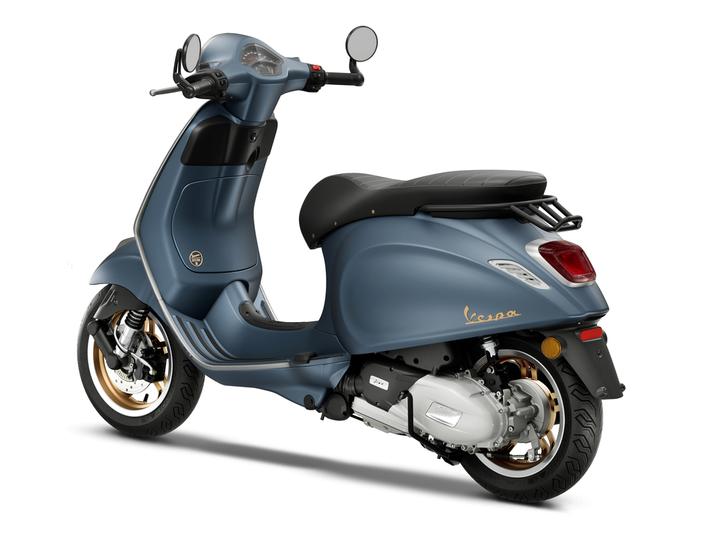A blue Vespa scooter viewed from the rear, showcasing its sleek design, black seat, and detailed rear light. The wheels have gold accents, and the handlebars include mirrors and a speedometer.