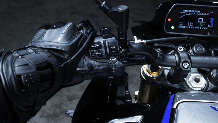 Close-up of a gloved hand on the handlebars of a motorcycle, interacting with control switches, including buttons for cruise control and modes, with a digital display in the background showing various metrics.