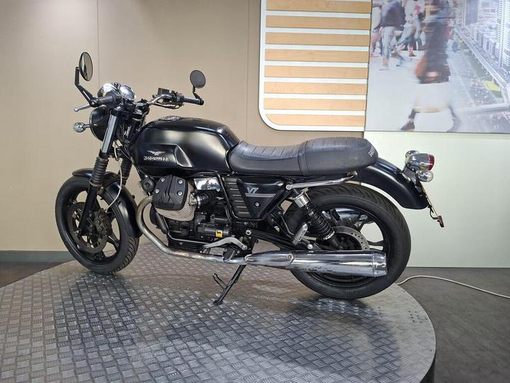 A sleek black motorcycle with a stylish design is showcased on a textured circular display platform against a minimalist background featuring a blurred image of people walking.