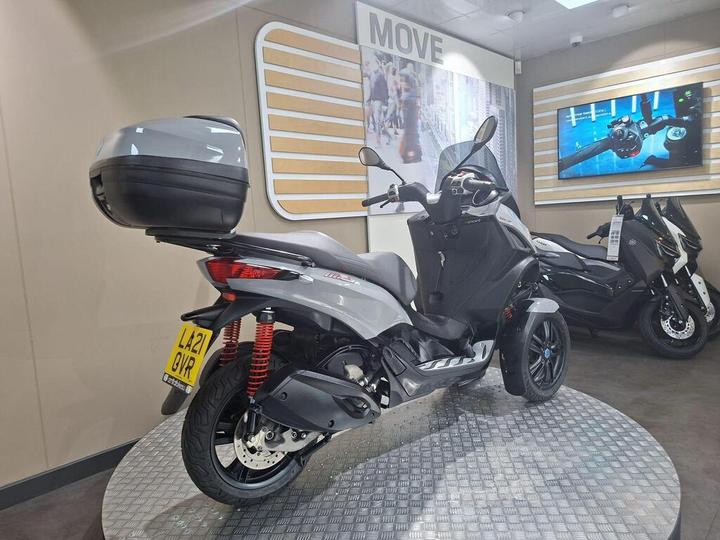 A grey scooter with a box on the rear, displayed on a checkered platform in a showroom setting, with a large image and another scooter visible in the background.