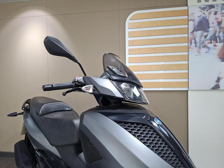 A close-up view of a gray scooter with a sleek design, showing the handlebars, mirrors, and headlight, set against a neutral background with a partially visible wall decor.