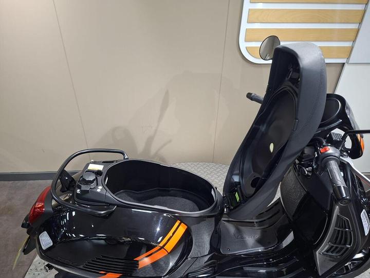 A close-up view of a black scooter with an open seat, revealing a storage compartment underneath. The scooter features an orange stripe design and is positioned against a neutral-colored wall.