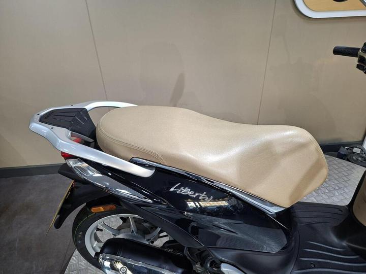A close-up view of a black scooter with a beige seat and silver rear rack, parked indoors.