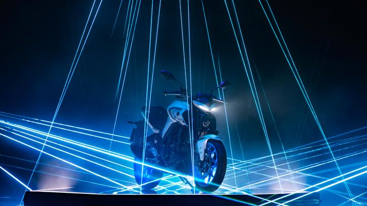 A white motorcycle is illuminated by blue laser beams in a dark environment, creating a dramatic effect.