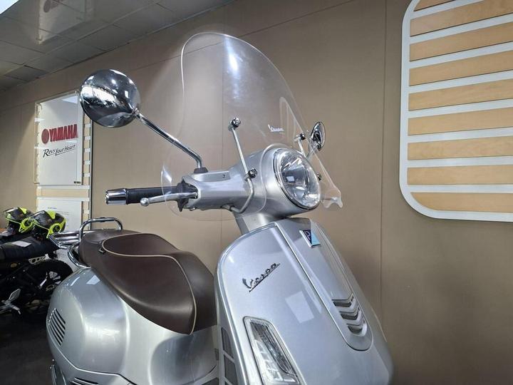 Close-up of a silver Vespa scooter with a clear windscreen, featuring chrome mirrors and a brown seat, displayed against a beige wall with a Yamaha logo in the background.