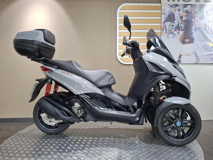 A gray three-wheeled scooter is displayed on a circular platform, featuring a black storage compartment at the rear, red shock absorbers, and a sleek design with prominent headlights and a windshield. The background includes a modern graphic display with people in a city setting.