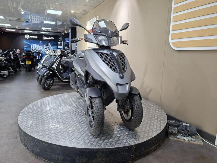 A gray three-wheeled scooter displayed on a turntable in a motorcycle showroom, with various other scooters in the background. The showroom features a sleek, modern interior.