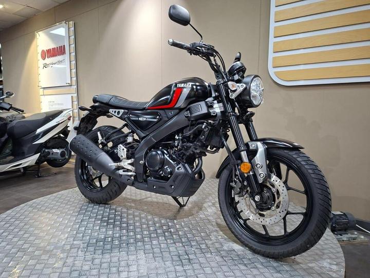 A Yamaha motorcycle is displayed on a textured floor against a neutral wall, featuring a black and red color scheme, circular headlight, and modern styling. A white scooter is partially visible in the background.