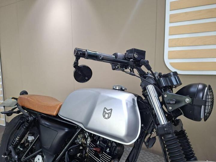 A close-up view of a custom motorcycle featuring a metallic silver fuel tank, vintage brown leather seat, and modern handlebars equipped with mirrors and a small camera. The backdrop consists of a minimalist wall with wooden accents.