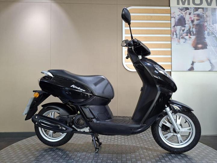 Peugeot Kisbee 50 2018 A sleek black scooter parked on a textured floor, featuring a comfortable seat, modern design, and white wheels, with a stylish background showcasing a blurred urban scene.