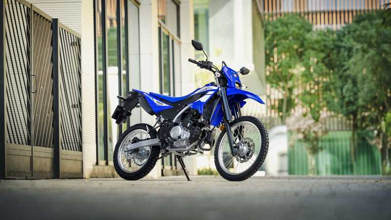 A blue dirt bike is parked on a smooth surface, positioned at an angle with a modern building and greenery in the background.