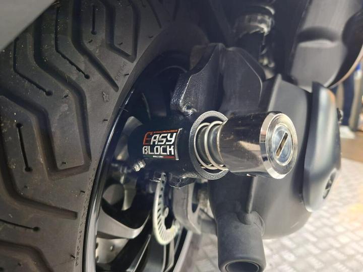 Close-up of a motorcycle wheel showcasing the "Easy Block" security lock attached to the axle, with visible tire tread patterns in the background.