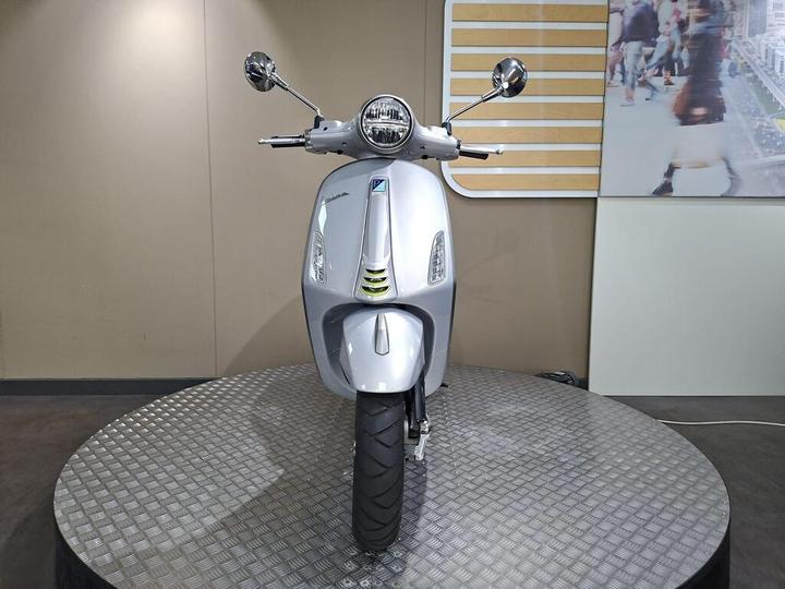 A silver scooter displayed on a circular platform with a textured surface, featuring a rounded front, chrome mirrors, and a modern design, against a neutral wall background.