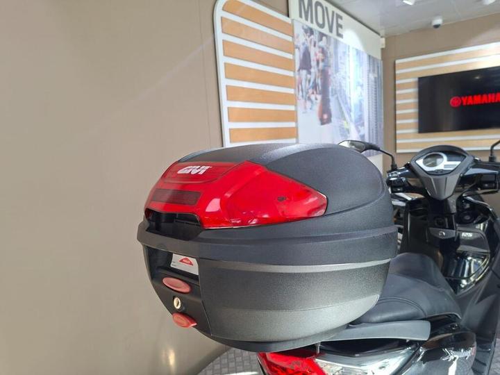 A close-up of a motorcycle's rear storage box, featuring a sleek black design with a red reflective top, mounted on the back of the motorcycle in a showroom setting.