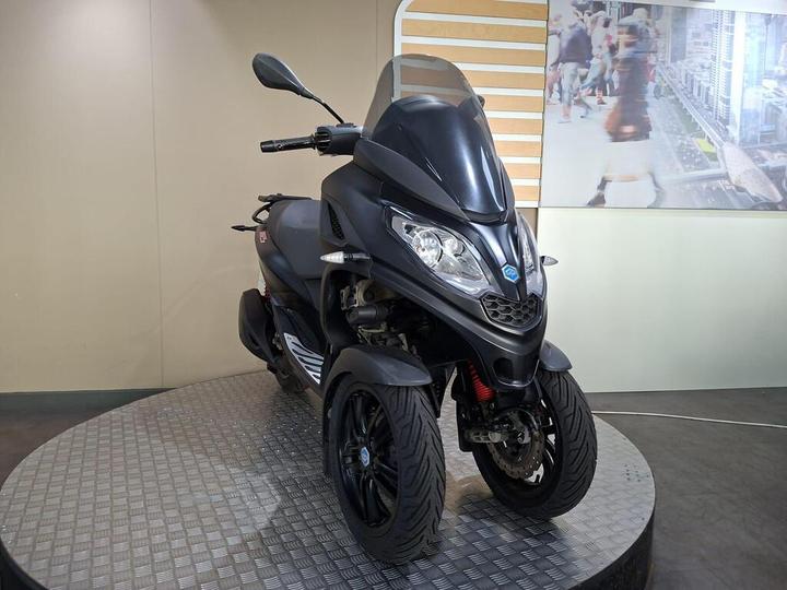 A black three-wheeled scooter displayed on a rotating platform against a neutral backdrop with a blurred cityscape image in the background.