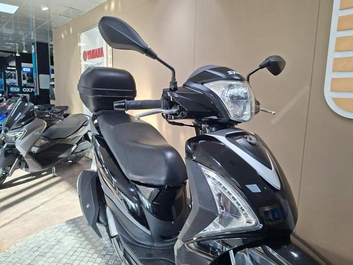 A black scooter is prominently displayed in a showroom, with its seat, handlebars, and rearview mirrors clearly visible. Other scooters can be seen in the background against a beige wall featuring the Yamaha logo.