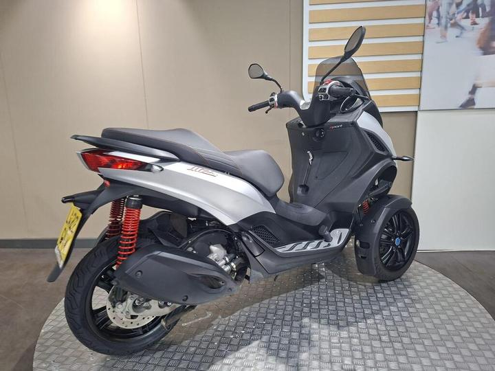 A three-wheeled scooter with a sleek silver body, red shock absorbers, and a black seat, displayed on a textured gray surface against a neutral background.