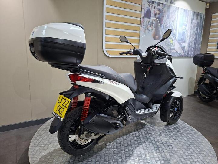 A stylish white and black scooter with a rear storage box, showcased on a circular metallic platform, in a modern indoor setting.