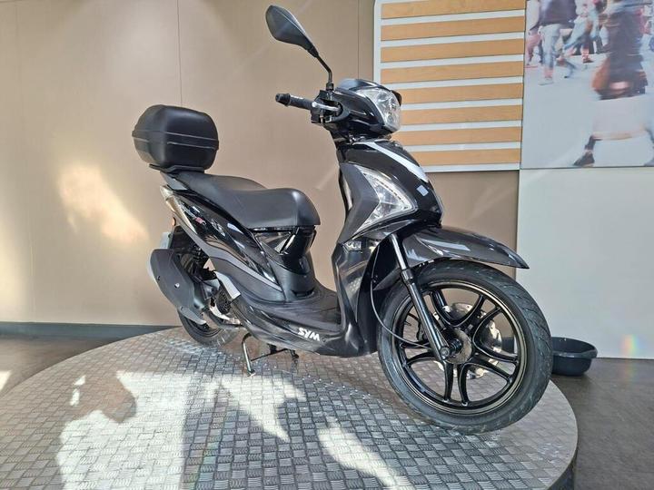 A sleek black scooter displayed on a circular patterned platform, featuring a stylish design, a comfortable seat, and a storage compartment on the rear. The background includes a light-colored wall and a framed image of people walking.