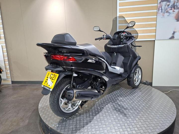 A black three-wheeled scooter is displayed on a circular stand in a showroom, showcasing its rear and side profile; a smooth floor and neutral-toned walls are in the background.