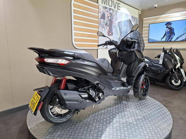 A black scooter displayed in a showroom, featuring red suspension springs and modern design elements, with another scooter visible in the background.