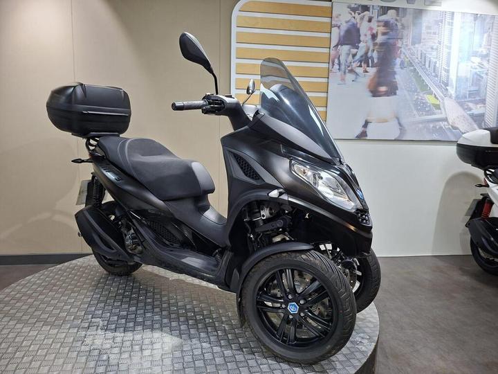 A black three-wheeled scooter with a prominent windshield and a rear luggage box, displayed on a textured circular platform against a backdrop featuring city scenes.