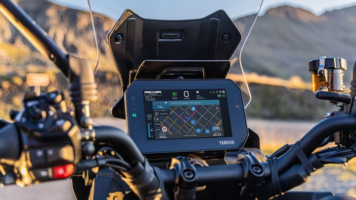 A close-up view of a motorcycle's dashboard featuring a large GPS screen displaying a street map, with navigation details visible. The background shows a mountainous landscape and part of the motorcycle's handlebar and controls.