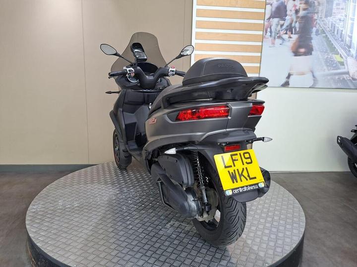 A gray three-wheeled scooter displayed on a rotating platform, featuring a rear view with a distinctive design, tail lights, and a license plate reading "LF19 WKL." The background shows a neutral wall with wooden elements.
