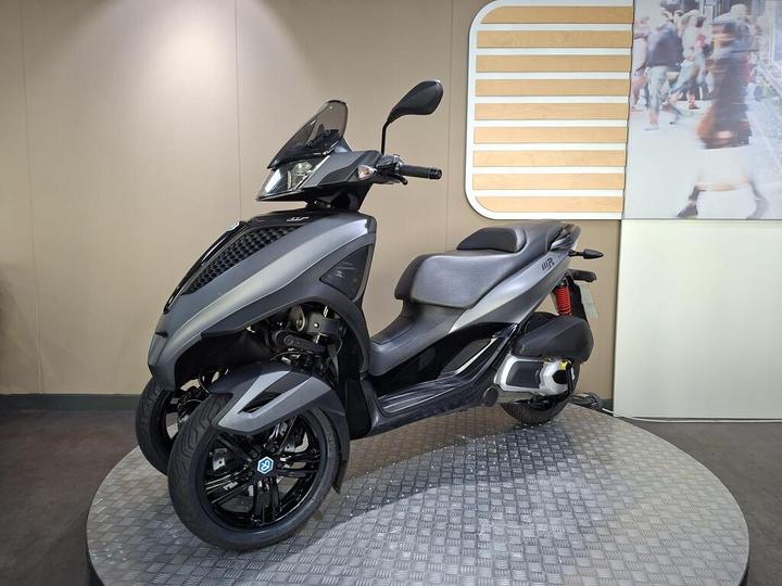 A modern gray three-wheeled scooter displayed on a circular platform with a textured metallic surface, against a neutral wall backdrop.