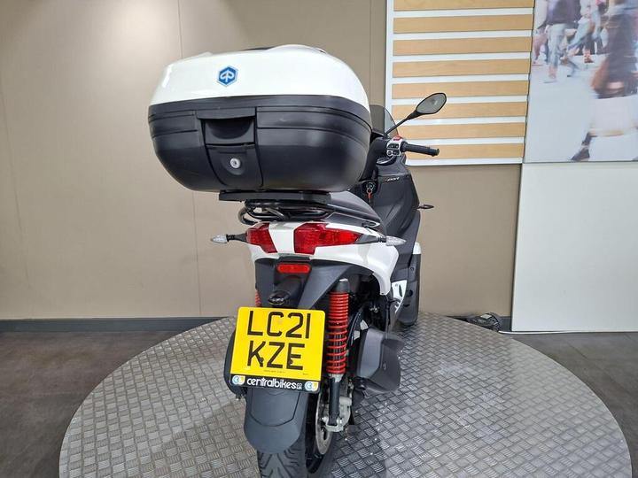 A rear view of a white scooter with a black top box, displaying the license plate "LC21 KZE," set against a neutral background with a circular platform.