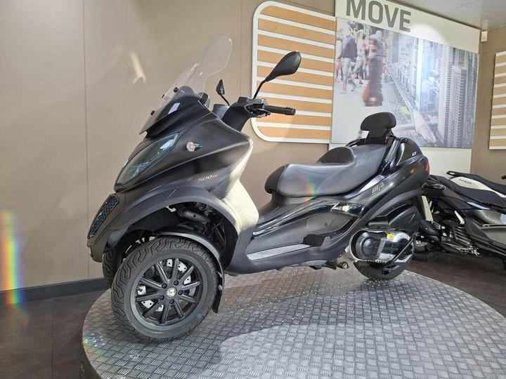 A black three-wheeled scooter displayed on a round platform in a modern showroom, with a large glass windshield and stylish design. In the background, there's a promotional wall featuring urban imagery and the word "MOVE."