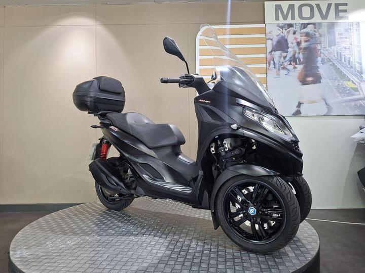 A sleek black three-wheeled scooter with a large windscreen and a cargo box on the rear, displayed on a circular platform against a neutral background.