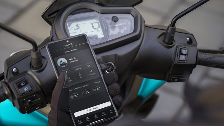 A close-up of a smartphone displaying a motorcycle app interface, alongside a motorcycle's handlebar. The app shows ride statistics, battery status, and current weather as a user prepares to ride.
