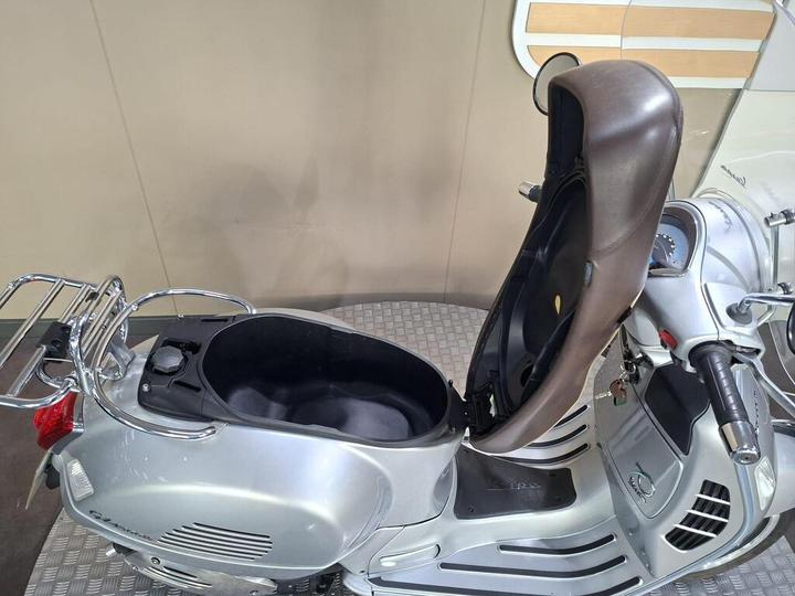 A close-up view of a silver scooter with a brown seat partially opened, revealing the storage compartment beneath. The scooter features a chrome handle, a speedometer on the front, and a textured floor. The background is a plain, light-colored wall.