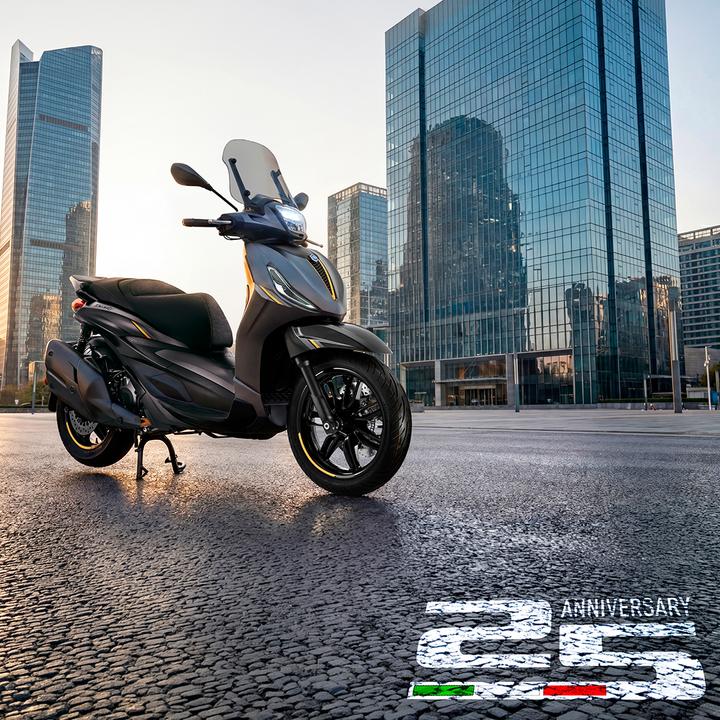 A sleek black scooter stands on a city street, framed by modern glass skyscrapers in the background, with a "25 ANNIVERSARY" graphic in the foreground.