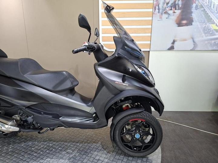 A black three-wheeled scooter is displayed, showcasing its sleek design and sporty features, including a windshield and modern wheels, against a neutral background.