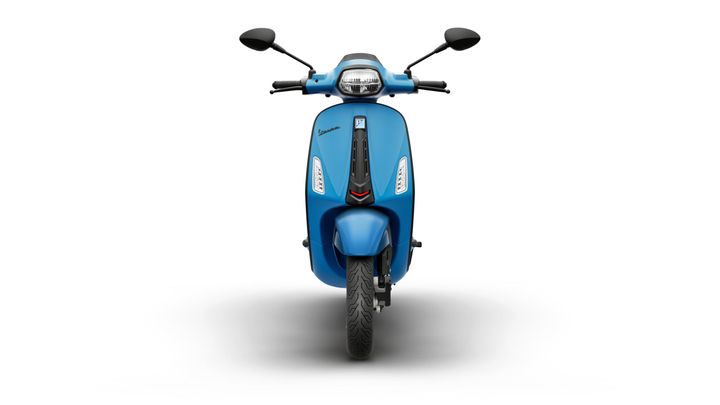 A blue scooter is facing forward, featuring a sleek design with a prominent front light and stylish detailing. The handlebars and mirrors are visible, with the rear wheel partially showing. The background is plain white, enhancing the scooter's appearance.