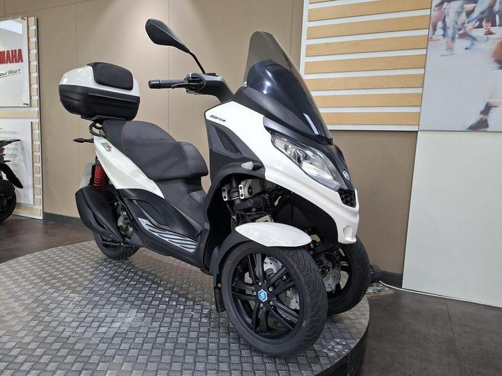 A white and black three-wheeled scooter displayed on a circular checkered platform, featuring a large windscreen, comfortable seating, and a rear storage compartment. The background includes a partial view of a showroom with promotional materials.