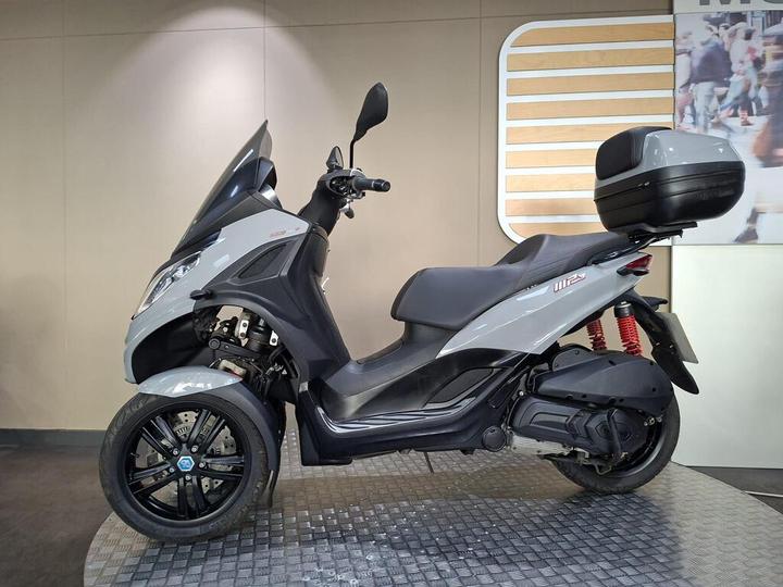 A side view of a gray three-wheeled scooter with a windshield, a spacious seat, and a rear storage compartment, displayed on a textured platform against a neutral backdrop.