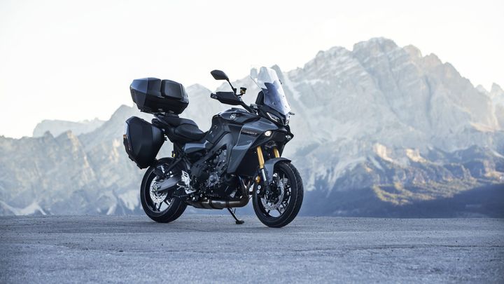 A sleek gray motorcycle parked on a scenic overlook with mountains in the background, featuring a tall windscreen and side panniers.