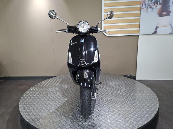 A black scooter is displayed facing forward on a circular platform, with bright chrome mirrors and an illuminated headlight, set against a neutral background.