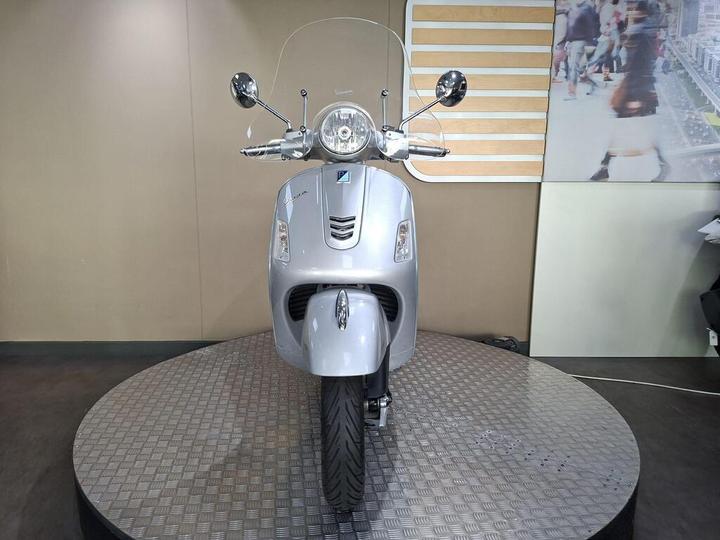 Front view of a silver Vespa scooter displayed on a circular platform, with a neutral background and visible reflections on the scooter's surface.