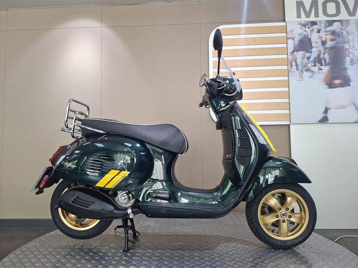 A green vintage scooter with gold wheels, a chrome rear rack, and yellow accents, displayed on a textured surface against a neutral background.