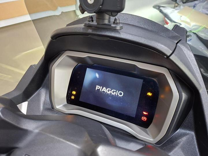 A close-up view of a Piaggio scooter dashboard display featuring a digital screen showing the "PIAGGIO" logo, accompanied by various indicator lights.