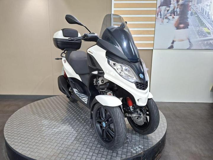 A white and black three-wheeled scooter displayed on a circular platform, featuring a transparent windshield, a storage compartment at the rear, and modern design elements. The background includes a blurred image of people walking.