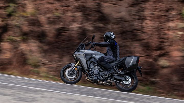 A rider in a black motorcycle outfit is cruising on a gray motorcycle along a winding road, surrounded by a blurred backdrop of reddish-brown terrain, conveying a sense of speed and motion.