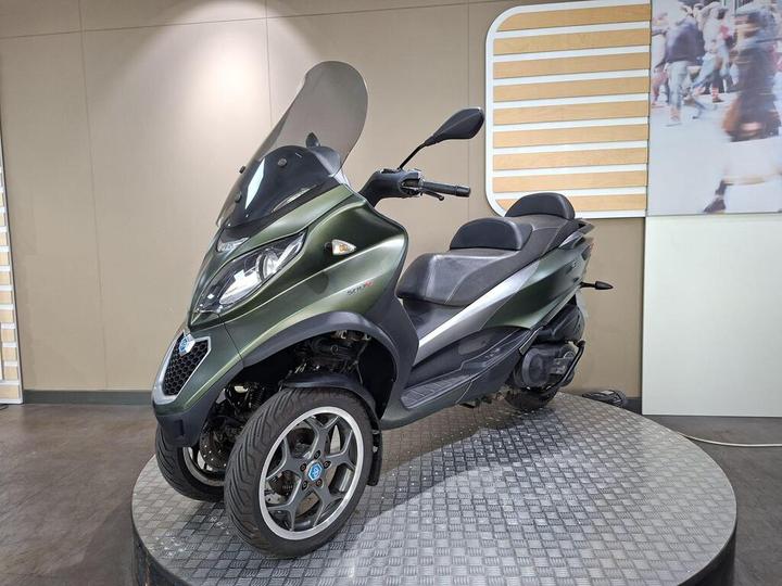 A green three-wheeled scooter displayed on a rotating platform, featuring a large windshield and a sleek design, set against a neutral background with subtle decorative elements.