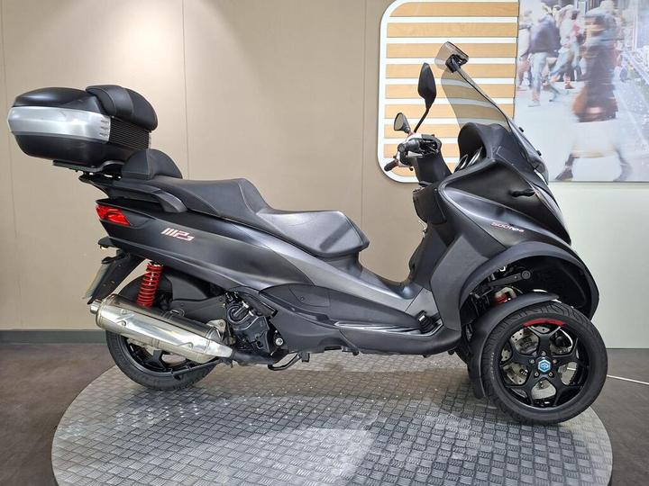 Piaggio MP3 500 HPE LT Sport 2020 A sleek black three-wheeled scooter with a large rear storage compartment, displayed on a circular platform with a textured surface. The scooter features sporty red rear suspension and distinctive wheel designs.