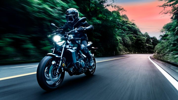 A motorcyclist in a black helmet and gear rides a sleek motorcycle on a winding road surrounded by lush greenery at sunset. The bike's headlights illuminate the path ahead as it accelerates.